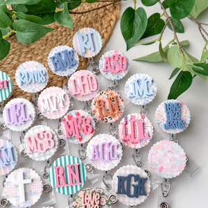 May include: A collection of round, decorative badge reels with various initials and phrases. The reels feature pastel colors, floral patterns, and scalloped edges. Each badge reel has a retractable cord and a clip for attaching to clothing.