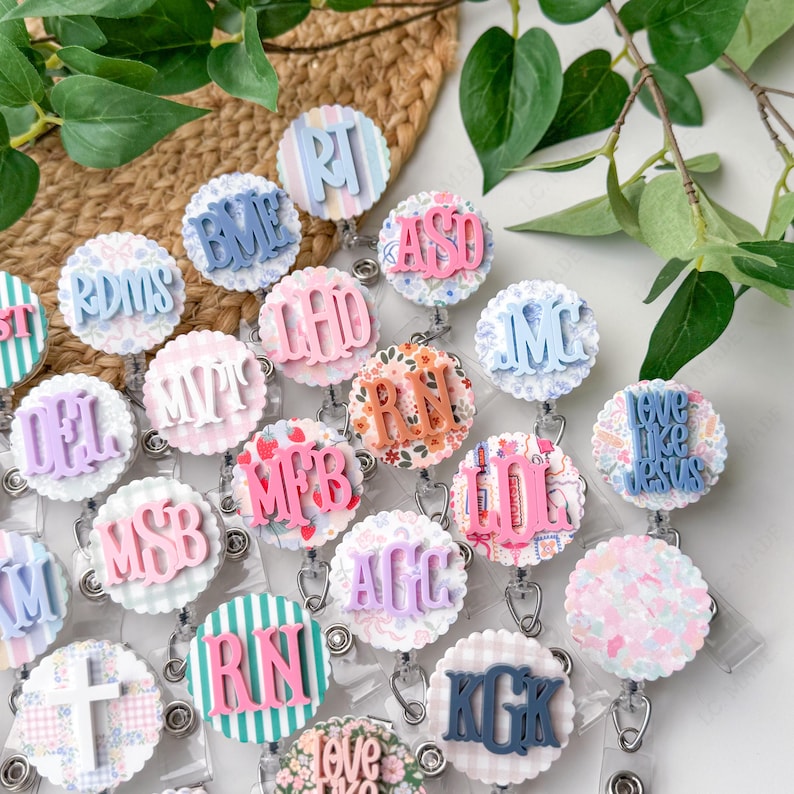May include: A collection of round, decorative badge reels with various initials and phrases. The reels feature pastel colors, floral patterns, and scalloped edges. Each badge reel has a retractable cord and a clip for attaching to clothing.
