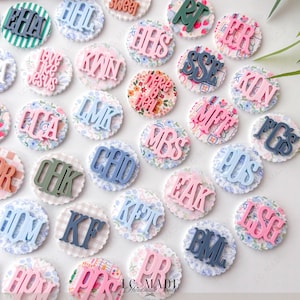 May include: A collection of decorative, personalized round embellishments. Each has a scalloped edge and a patterned fabric base. The embellishments feature various initials and phrases in shades of blue, pink, and green. The text "LC MADE Handmade Foods" is visible.