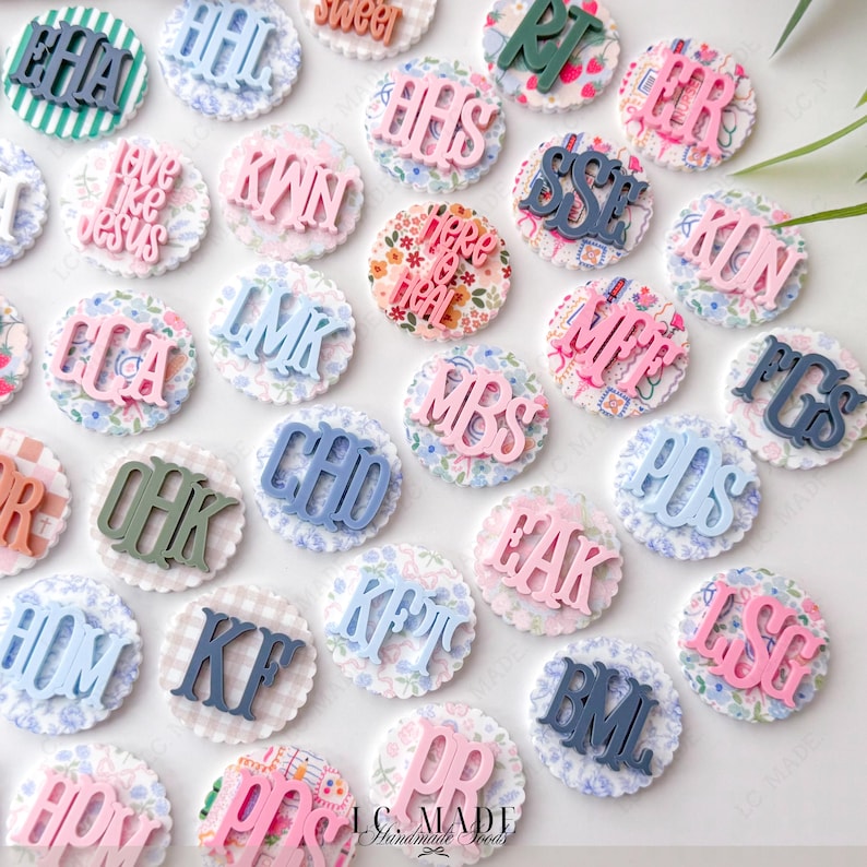 May include: A collection of decorative, personalized round embellishments. Each has a scalloped edge and a patterned fabric base. The embellishments feature various initials and phrases in shades of blue, pink, and green. The text "LC MADE Handmade Foods" is visible.