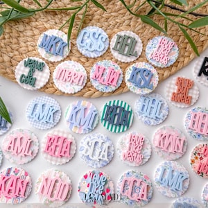 May include: A collection of round, scalloped-edge embellishments with various initials and phrases in pastel colors. Some have floral or gingham patterns, while others feature striped or solid backgrounds. The text includes "Jesus is Cool" and "Love Like Jesus."