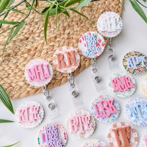 May include: A collection of round badge reels with various designs and initials. The reels feature floral, plaid, and patterned backgrounds with raised lettering in pink, peach, and white. Some reels have the text "love like jesus".