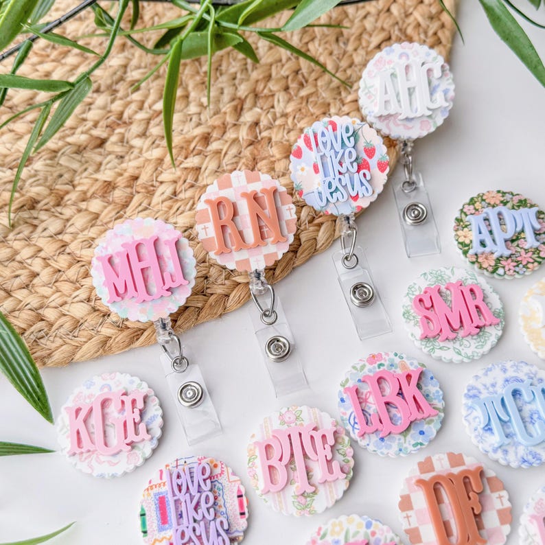May include: A collection of round badge reels with various designs and initials. The reels feature floral, plaid, and patterned backgrounds with raised lettering in pink, peach, and white. Some reels have the text "love like jesus".