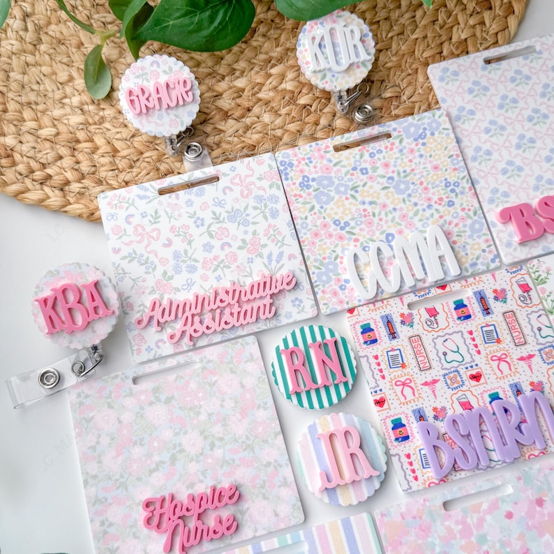 May include: A collection of floral-patterned badge reels and name tags. The tags feature names and titles like "Grace", "Administrative Assistant", and "Hospice Nurse" in pink and white. The background includes a woven basket and greenery.
