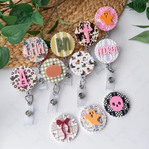 May include: A collection of circular badge reels featuring various designs including a pink letter A on leopard print, a pumpkin on green plaid, a pink cross on cow print, a ghost on pink, and a pink skull on a black and white checkered pattern.