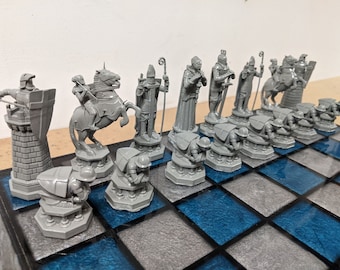 May include: A full grey chess set featuring intricately designed medieval figures, such as knights on horseback, bishops, and pawns. The chess pieces are set out on a blue and grey chequered board, prepared for a game.