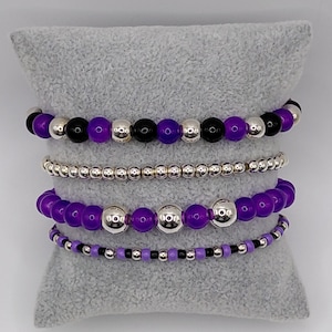 Könnte beinhalten: Vier Armbänder mit Perlen in Lila, Schwarz und Silber. Die Armbänder werden auf einem grauen Kissen präsentiert. Die Perlen variieren in Größe und Material und erzeugen einen geschichteten Effekt. Die Armbänder sind eine Mischung aus runden und kleinen Perlen.