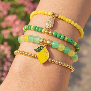 May include: Four beaded bracelets in yellow, green, and gold. One bracelet features a gold fish charm, and another has a yellow lemon charm with a green leaf. The bracelets are worn on a wrist.