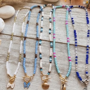 May include: Five beaded necklaces featuring various color schemes including white, light blue, turquoise, and dark blue. Each necklace is finished with a gold-toned toggle clasp and a unique charm, such as a butterfly, a seashell, or a conch shell.