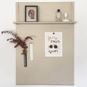 May include: A beige metal wall-mounted organizer featuring a top shelf holding a framed abstract portrait, a clear glass bottle, and a white candle holder. The board displays a print with the text "je ne sais quoi" and sunglasses, plus two glass vases.