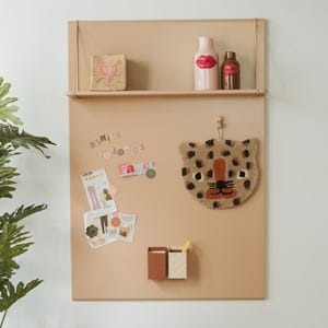 Può includere: Un organizer beige da parete con un ripiano contenente un cuore intrecciato, un vaso rosa a forma di labbra e una bottiglia marrone. Sotto, un arazzo da parete a forma di testa di leopardo, magneti e due piccoli contenitori sono attaccati.