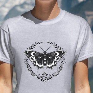 May include: A light gray t-shirt featuring a black and white butterfly design encircled by a wreath of leaves. The butterfly has detailed wing patterns. The shirt is made of a soft material and is suitable for casual wear.