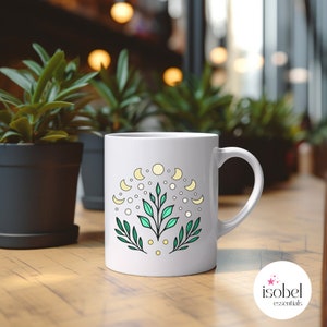 May include: White ceramic mug featuring a celestial design with moons, dots, and leaves in yellow, green, and white. The mug has a curved handle and is set on a wooden surface.