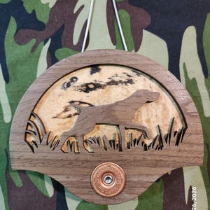 May include: A semi-circular wooden wall hanging featuring a laser-cut silhouette of a hunting dog in tall grass against a light wood background. A copper-coloured Remington Peters shotgun shell casing is centred at the bottom with a metal hanger.