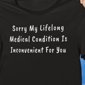 以下が含まれることがあります： 「Sorry My Lifelong Medical Condition Is Inconvenient For You」の白文字がプリントされた黒いTシャツ。青い空と雲を背景に、Tシャツが掲げられています。