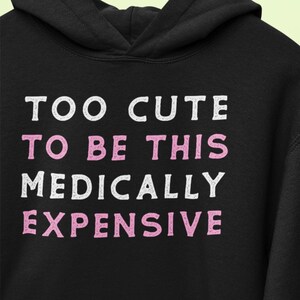 Może przedstawiać: Czarna bluza z kapturem z napisem "TOO CUTE TO BE THIS MEDICALLY EXPENSIVE" w białych i różowych literach. Bluza wisi na drewnianym wieszaku na jasnym zielonym tle.