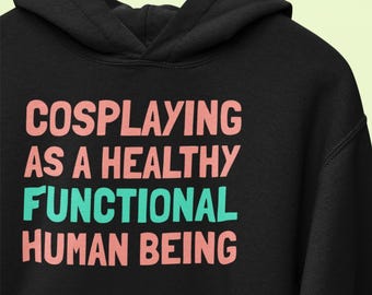 May include: Black hooded sweatshirt with the text "COSPLAYING AS A HEALTHY FUNCTIONAL HUMAN BEING" in pink and teal lettering. The sweatshirt is hanging on a wooden hanger against a light green background.
