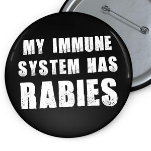 May include: A round black pin-back button featuring the white distressed text MY IMMUNE SYSTEM HAS RABIES. The button has a metallic silver back with a horizontal pin mechanism visible, showing the construction of the circular accessory.