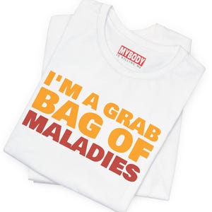 Op de afbeelding: Wit T-shirt met de tekst "I'M A GRAB BAG OF MALADIES" in oranje en rood. Het shirt heeft ook de tekst "MYBODY IS KILLING ME" in een rode rechthoek. Het T-shirt is opgevouwen.