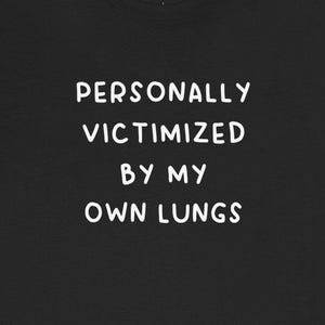 May include: Black t-shirt with white text that reads "PERSONALLY VICTIMIZED BY MY OWN LUNGS".