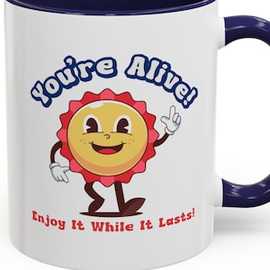 May include: White ceramic coffee mug with a blue rim and handle. The mug features a cartoon sun with a smiling face and the text "You're Alive! Enjoy It While It Lasts!"