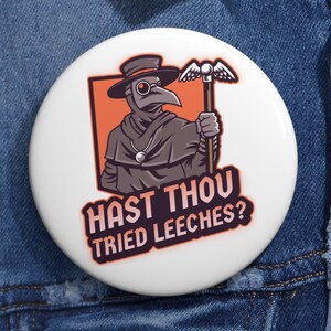 May include: A white button with a cartoon image of a plague doctor wearing a black robe and a long beak mask. The doctor is holding a staff with a bird on top. The text "Hast thou tried leeches?" is written in orange and black on the button.