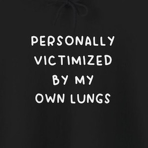 May include: Black hoodie with white text that reads "PERSONALLY VICTIMIZED BY MY OWN LUNGS".