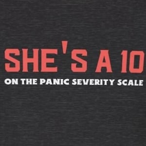 Puede incluir: Una camiseta gris oscuro con el texto "SHE'S A 10 ON THE PANIC SEVERITY SCALE" en letras rojas.