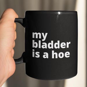 Może przedstawiać: Czarny ceramiczny kubek z białym napisem "my bladder is a hoe". Kubek ma czarny uchwyt i jest trzymany przez dłoń. Kubek jest cylindryczny i wydaje się być standardowego rozmiaru.