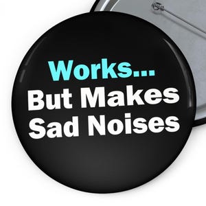 Könnte beinhalten: Ein schwarzer Button mit dem Text "Works... But Makes Sad Noises" in Weiß und Hellblau. Der Button hat eine Metallnadel auf der Rückseite.