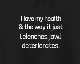 May include: Black hoodie with white text: "I love my health & the way it just [clenches jaw] deteriorates." The text is centered and in a sans-serif font. The hoodie's drawstring is visible at the top.