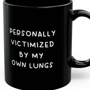 May include: Black ceramic coffee mug with white text that reads "PERSONALLY VICTIMIZED BY MY OWN LUNGS".