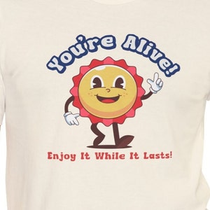 May include: Off-white t-shirt with a cartoon bottle cap character. The character has a smiling face, red and yellow accents, and the text "You're Alive! Enjoy It While It Lasts!" in blue and red.