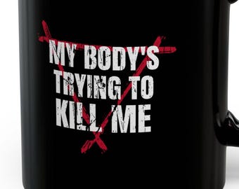 May include: Black ceramic mug with the text "MY BODY'S TRYING TO KILL ME" in white, crossed out with red lines. The mug has a black handle and a glossy finish.