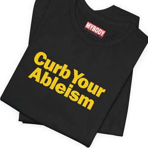 May include: Black t-shirt with the text "Curb Your Ableism" in yellow letters. The text "My Body Is Killing Me" is printed on a red tag attached to the shirt.