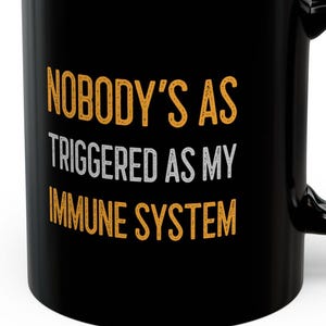 Może przedstawiać: Czarny kubek do kawy z napisem "Nobody's as triggered as my immune system" w pomarańczowych i białych literach.