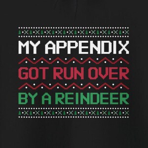 Puede incluir: Suéter navideño negro con un diseño blanco, rojo y verde. El texto en el suéter dice "My appendix got run over by a reindeer."