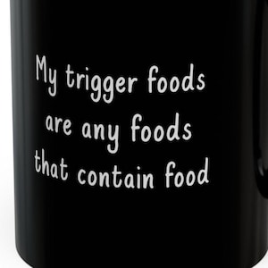 Può includere: Tazza da caffè nera con testo bianco che recita "I miei cibi trigger sono tutti i cibi che contengono cibo".