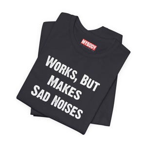 May include: Black t-shirt with the text "WORKS, BUT MAKES SAD NOISES" in white block letters. The shirt has a crew neck and is folded. The brand label "MYBODY IS KILLING ME" is visible.