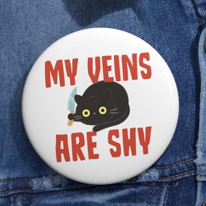 May include: A white button with the text "MY VEINS ARE SHY" in red. A black cat with yellow eyes is holding a small knife. The button is round and has a glossy finish.