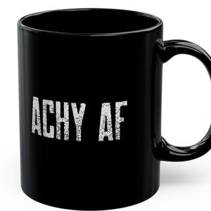 Puede incluir: Taza de café negra con el texto "Achy AF" en letras blancas.
