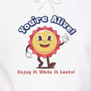 May include: White hoodie featuring a cartoon sun character wearing a red and white cap. The sun is smiling and has a white glove on its right arm. The text "You're Alive!" is in a blue circle above the sun. The text "Enjoy It While It Lasts!" is below the sun.