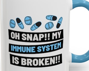 May include: White ceramic mug with a blue rim and handle. The mug features a design of blue pills with a black outline and the text "OH SNAP!! MY IMMUNE SYSTEM IS BROKEN!!"