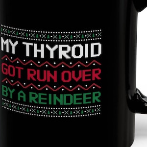 Puede incluir: Taza de café negra con un diseño blanco, rojo y verde que dice "My Thyroid Got Run Over By A Reindeer".