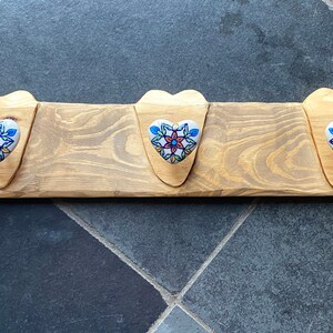 Pode incluir: Um cabide de parede de madeira com três ganchos em forma de coração. Cada gancho apresenta um botão de cerâmica com um design floral azul, vermelho e branco. A madeira tem um acabamento natural.