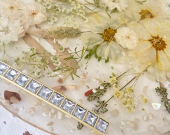 May include: A round, resin tray with embedded dried flowers in shades of white and yellow. A gold-toned bar with square, clear crystals is placed on the tray. The tray is decorated with small, white pebbles.