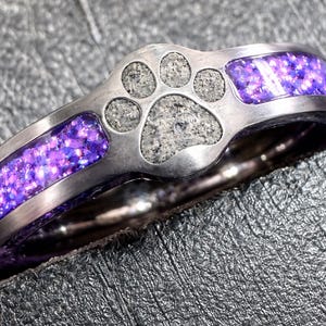 May include: A silver ring with a paw print design. The ring features a central paw print made of a gray stone, flanked by sections filled with sparkling purple material. The ring is set against a textured gray surface.