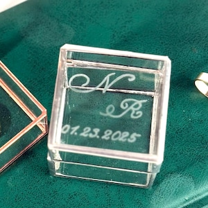 Op de afbeelding: Twee doorzichtige ringendoosjes met gouden ringen op een donkergroen oppervlak. Eén doosje is open en onthult een gouden ring binnenin. Het andere doosje is gesloten en heeft initialen en een datum op de voorkant gegraveerd. Het open doosje heeft een roségouden frame.