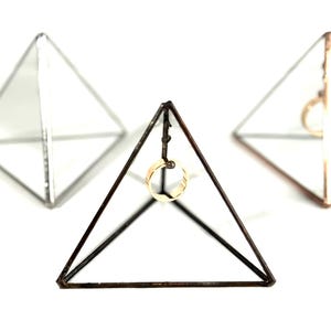 Puede incluir: Tres expositores geométricos de vidrio en forma de pirámide. El expositor central es de color bronce, con un anillo dorado suspendido en su interior. Los otros dos expositores son de color plateado y cobrizo, cada uno con un anillo.