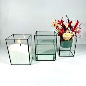 May include: Three square glass terrariums with black frames. The terrarium on the left contains a white candle. The middle terrarium is empty. The terrarium on the right contains dried flowers in shades of red, white, and cream.